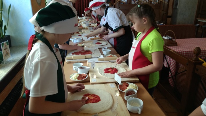 Children are spreading tomato sauce on pizza dough at Mama Roma's.