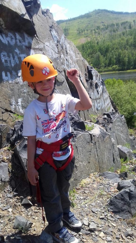 boy poses in climbing helmet and harness in green hilly setting with cliffs